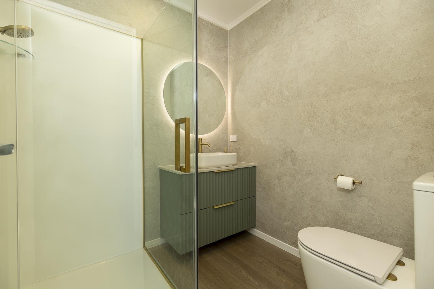 The renovated bathroom: large-format stone-look tiles, a fluted green vanity, round backlit mirror and brass fittings.